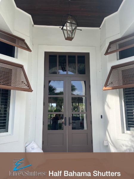 Double brown bahama shutters installed on an off-white stucco home with a dark brown door in Sanibel FL