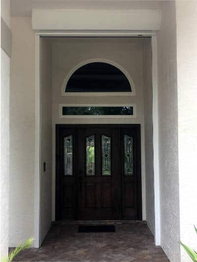View of the doorway of the home. The image shows two custom, impact windows above the home's entry door. One window is an arch, and the other is a skinny, rectangle shape. They both have white vinyl frames. Windows installed on a home in Bonita Springs Florida.