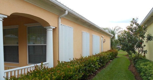 White accordion shutters shown installed over the windows on the side of a yellow house