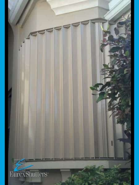 Beige colored accordions shown closed over the front window of a beige home.