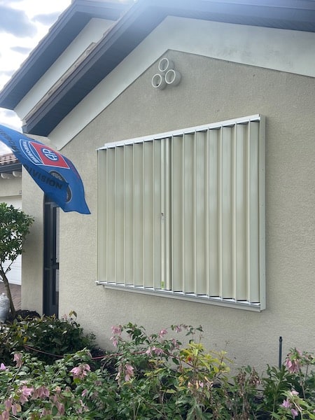 Beige accordion shutters with locks on a beige home. Shown in the closed position.