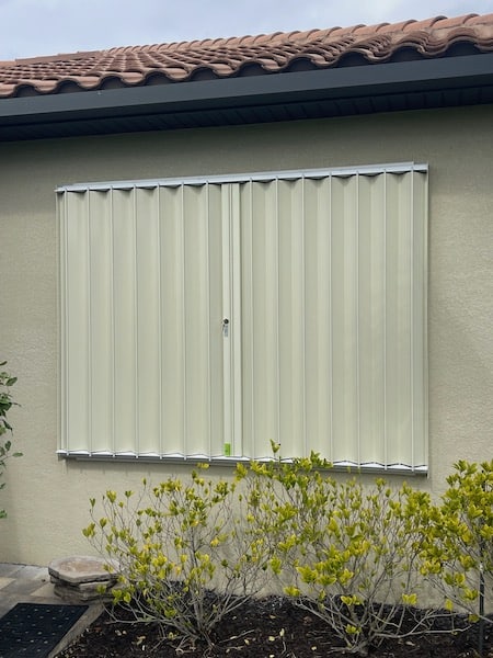 Beige accordion hurricane shutters with locks on a beige home. Shown in the closed position.