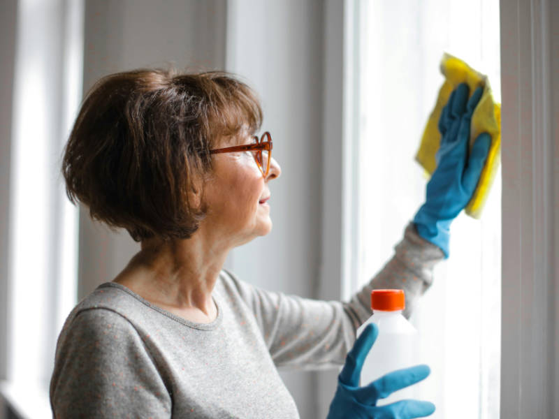 Woman with a grey shirt and blue rubber gloves cleaning inside of her windows with a microfiber cloth.